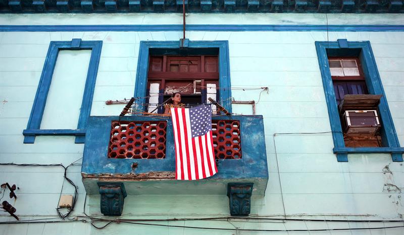 Una mujer se asoma a un balcón, junto a una bandera de Estados Unidos, hoy en una calle de La Habana
