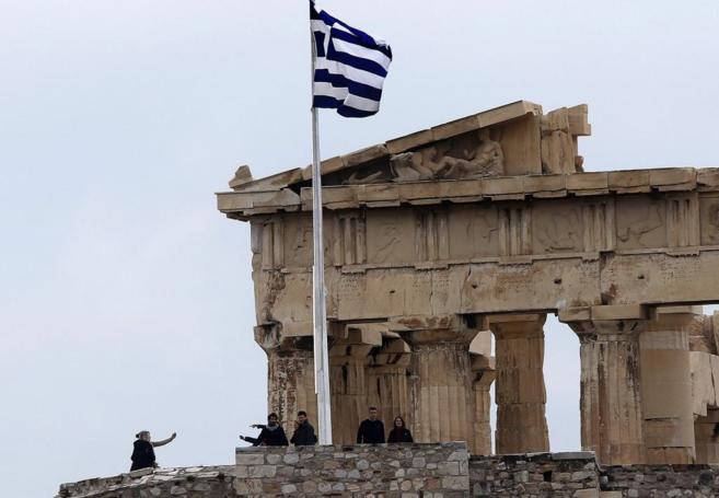 Una bandera griega ondea sobre la Acrópolis mientras turistas visitan las ruinas arqueológicas
