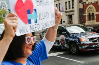 Un automóvil con la cara de Bernie Sanders pasa junto a una simpatizante de Hillary Clinton en Broad Street, Filadelfia, en Pensilvania
