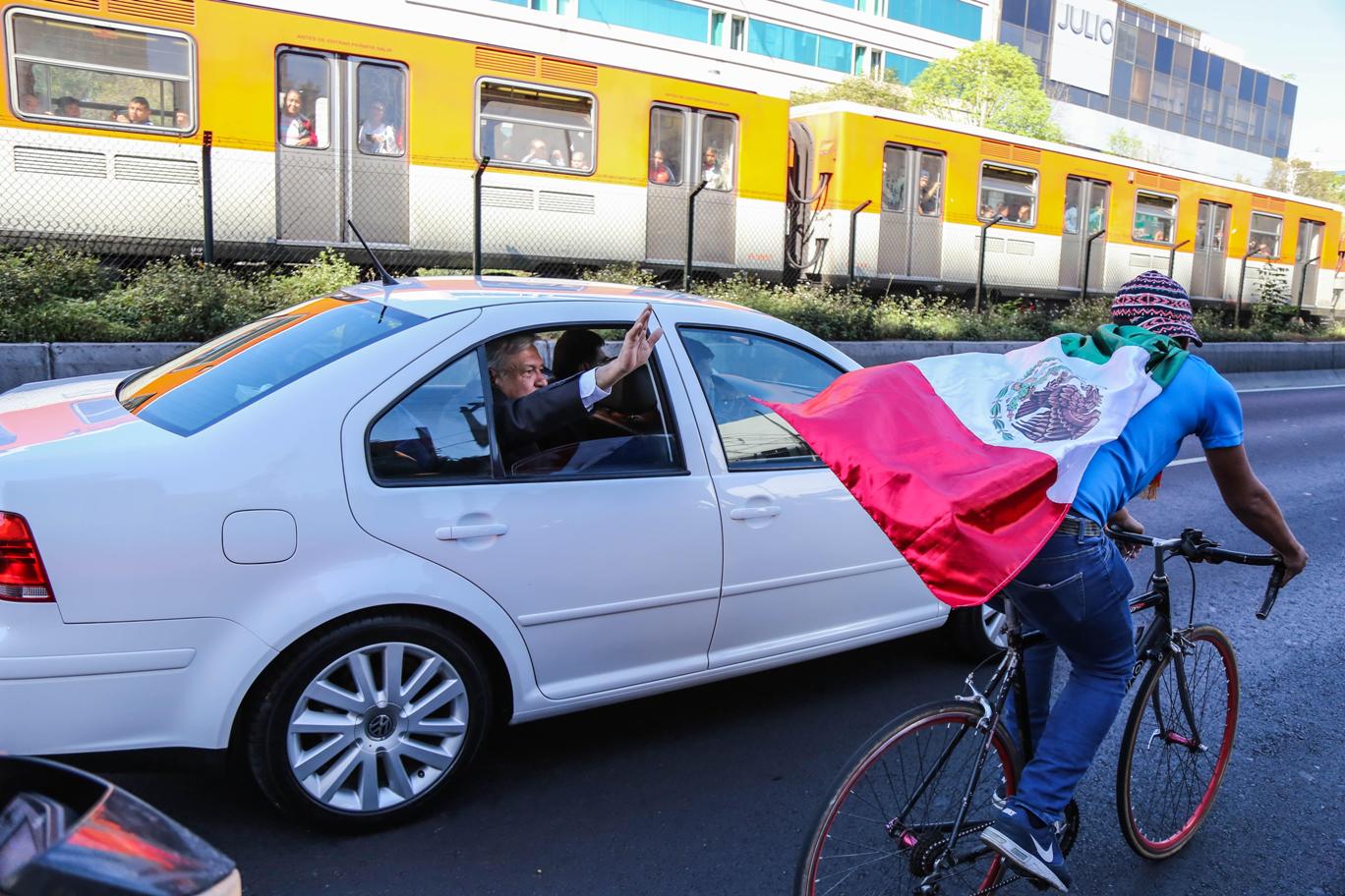 AMLO estira el brazo por la ventanilla para saludar a un ciclista que va a su costado con una bandera de México atada al cuello y que ondea sobre su e