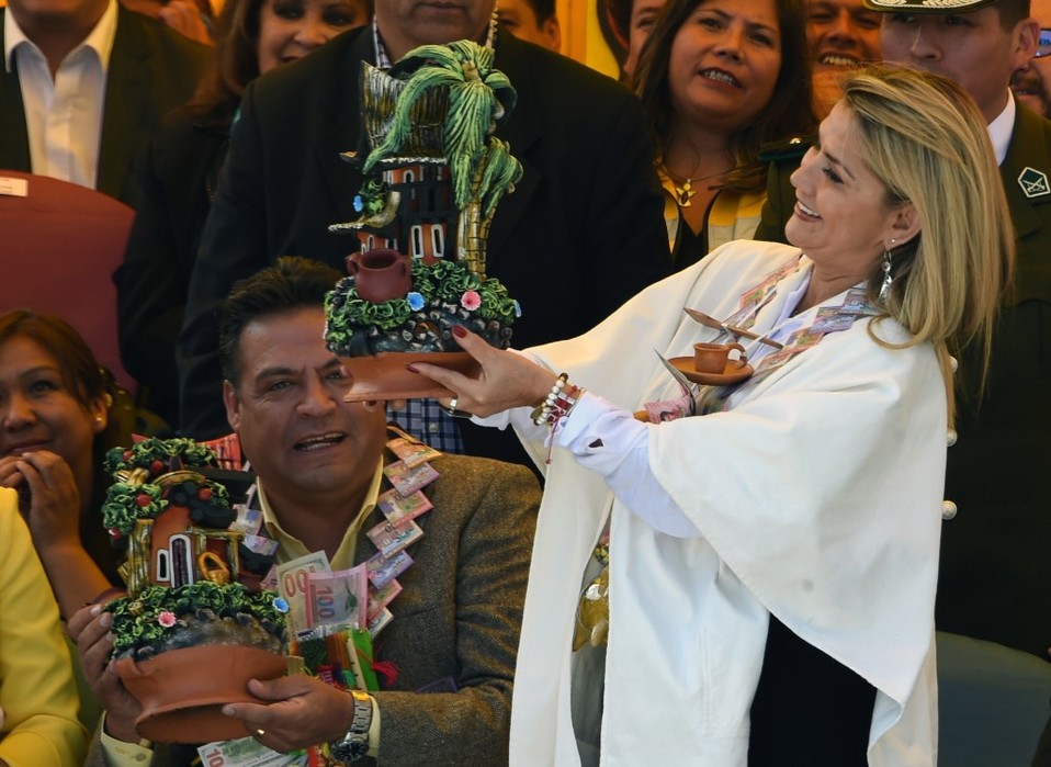 Jeanine Áñez sostiene un hogar en miniatura y lleva un collar hecho de dinero falso durante la inauguración de la feria de la Alasita. (AFP)