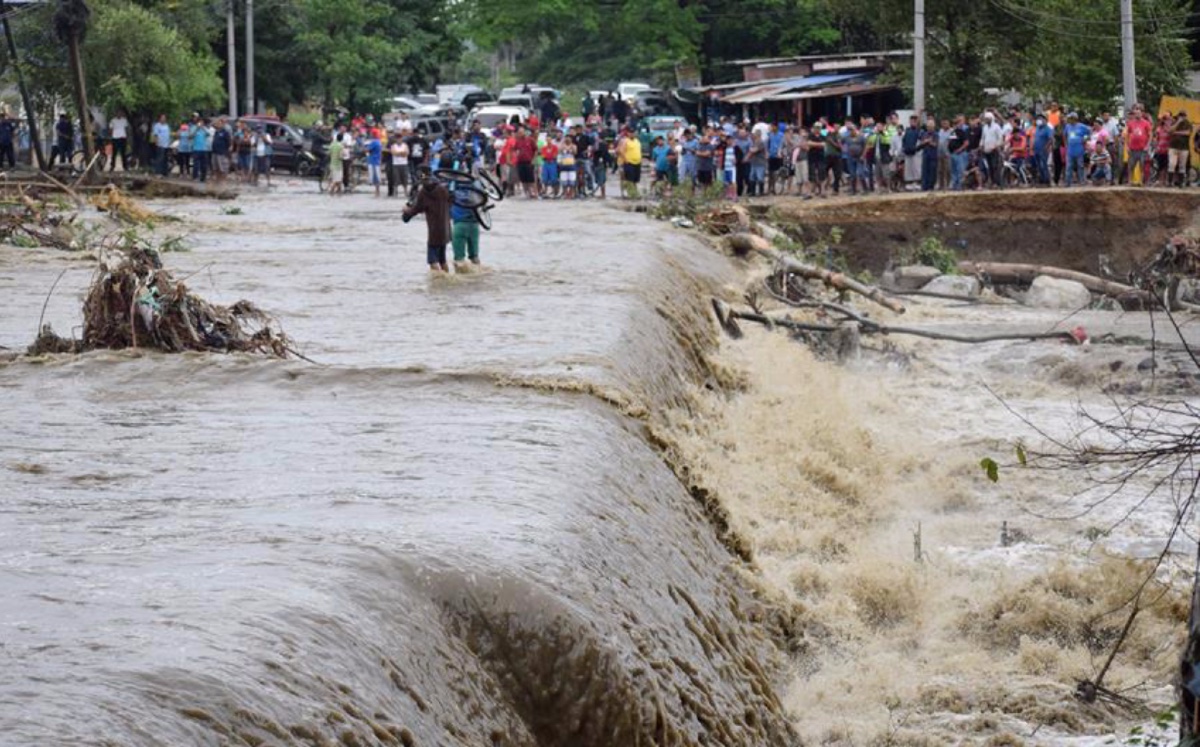 Decenas de personas observan a dos hombres cruzar una acaudalada inundación sobre una vía destruida por el desbordamiento del río Chamelecón. (EFE)