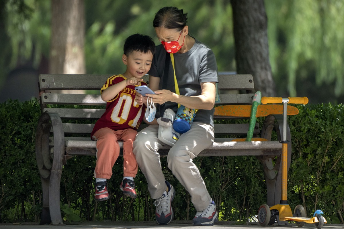 Una mujer con mascarilla y un niño observan un celular, sentados en una banca en un parque público en Beijing. (AP)