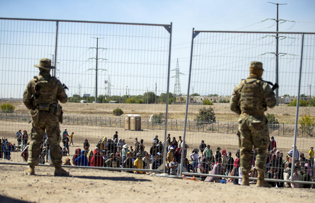 Varios migrantes esperan junto a la cerca fronteriza ante la mirada de guardias nacionales de Texas