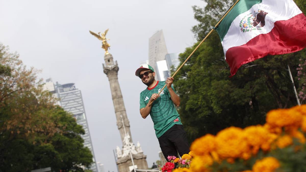 Un aficionado sonriente de la selección mexicana ondea la bandera de México frente al Ángel de la Independencia en Ciudad de México, mostrando su apoyo temprano para el partido inaugural.
