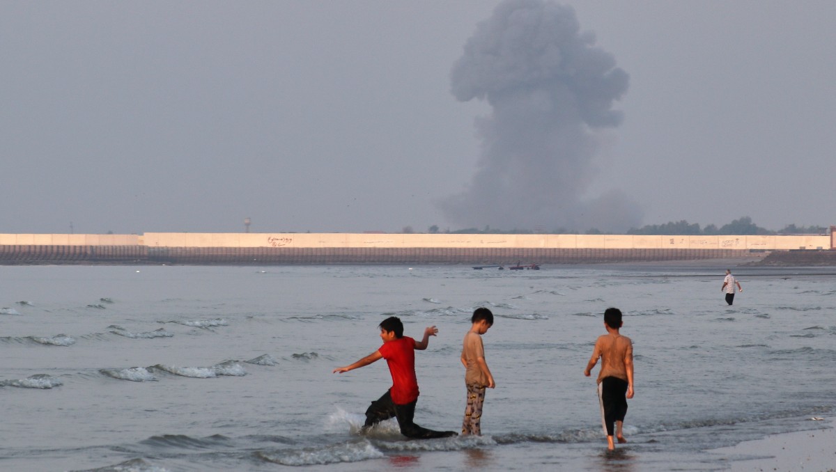 Tres niños juegan en el agua en el estrecho de Ormuz mientras al fondo se alza una columna de humo de una explosión, cerca de Bandar Abbas, Irán. | AP